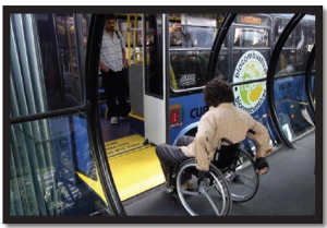 A person in a manual wheelchair is entering onto the short yellow ramp into the bus from the tube shaped bus shelter