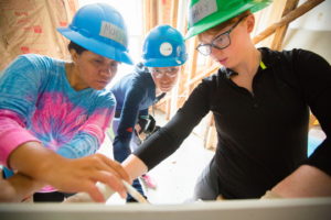 Picture of three young women wearing hard hats and holding pens and looking at a drawing on a table top