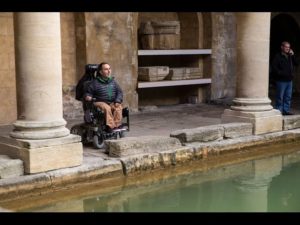 man in a powered wheelchair looking out between Roman columns and across the water in the Roman Bath