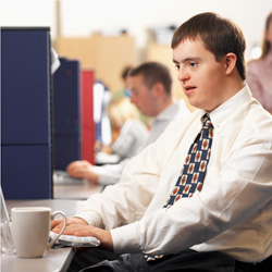 a young man with Down syndrome sits at a computer workstation. He is wearing a white shirt and patterned tie. there are other workers at workstations in the background. Down Syndrome and building design.