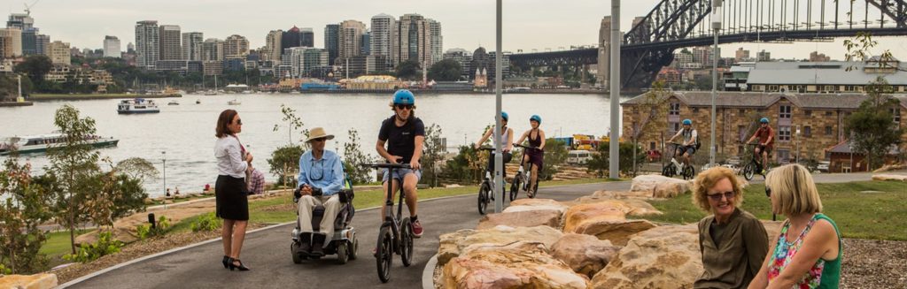Landscape view of Barangaroo Parkland showing a pedestrian, wheelchair user, cyclist and pram pusher. Mentioned in EU tourist guide.