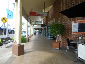 Wide footpath in a shopping strip which has a veranda overhead. There are planter boxes and a seat.