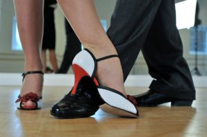 The feet of two dancers. The woman is wearing red and white shoes and the man regular black shoes