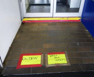 A yellow caution sign is taped to the ground with red tape. The doorway entrance has a step below the door with yellow and red tape on it.