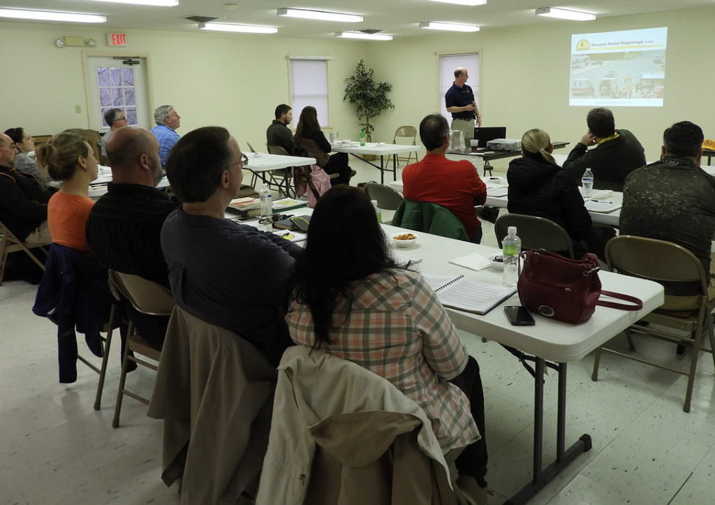 Adults seated at tables in a classroom setting looking forward to the instructor at the front of the room