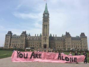 A group of people standing holding a pink banner with the words You are Not Alone, but you can't see the word NOT because it is in pale red and blends into the background colour