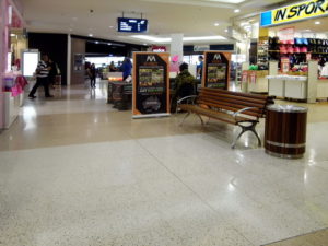 Picture of a shopping mall with a plain grey floor and shops on each side. Thre is a woodend bench with armrests and backrest. In the distance you can see more shops.