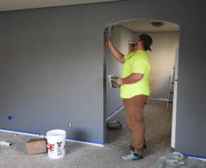 A man in a bright yellow T shirt is painting and archway in a wall inside a home. The wall is grey and there are tools on the floor.