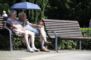 An older man and woman sit on a wooden slatted park bench. The man is holding a blue umbrella to shade from the sun. There is another empty bench next to them. They are sitting alongside the path and there are trees behind them.