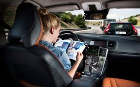 A woman with blonde hair sits reading a magazine resting on the steering wheel of the car she is (not) driving. The road and other cars are visible through the windscreen as the photo was taken from the back seat.