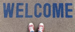 A mid grey doormat viewed from above with the word welcome in blue. At the bottom of the picture you can see the tops of a pair of red and white sneakers. Creating inclusion for everyone.