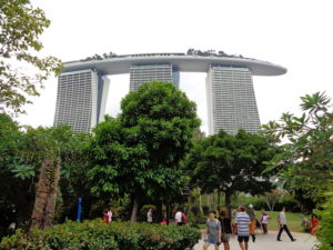 A distance picture of a three column building in Singapore with trees and people in the foreground. Sustainability and universal design.