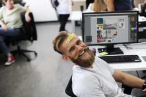 A bearded man in a white shirt leans back from his desk and computer. He is laughing and has a sticker on his forehead that reads, be happy.