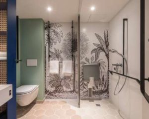 Hotel bathroom with walls decorated with line drawings of palm trees and other plants. There is a shower seat, hand held shower and a toilet pan that only allows for side transfer.