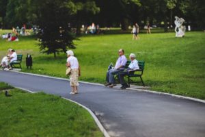 An older woman walks on a bitumen path in a park. Two older men are sitting on a seat along the pathway.