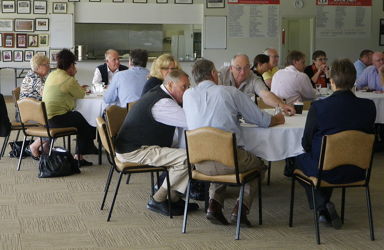 Older people sit at round tables discussing questions. There are four round tables shown in this picture.
