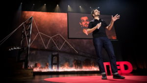 Johan Carey in jeans and black polo shirt is on the stage at a Ted Talk
