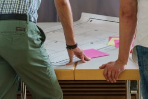 A desk with a large sheet of paper and pink post it notes. A person stands with their hand resting on the table. Inclusive design is more than a checklist.