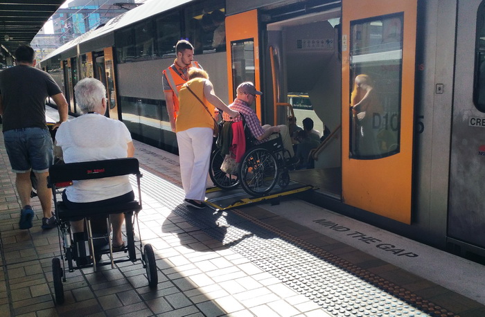 A woman is pushing a man in a wheelchair up a ramp into the train. The train guard looks on. Another woman in a wheelchair waits for her turn. A man with a stroller is also in the picture.