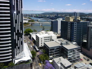 View from high building in Brisbane overlooking building roofs and the Brisbane river and bridges. Jacaranda trees can be seen in the street.