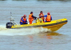 Four men with orange lifejackets are standing in a yellow State Emergency Service boat on a swollen river.