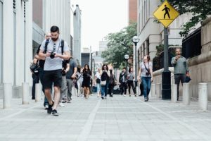 Pedestrians are walking towards the camera. They are on a wide walkway. Some people are looking at their phones. They are dressed for warm weather. There are buildings on each side of the walkway