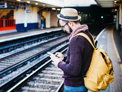 A man stands on a train platform looking at his smartphone. He is wearing a hat and has a bright yellow backpack.