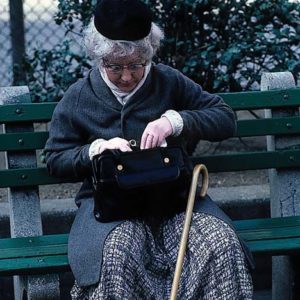 Patricia Moore sits on a park bench looking in her handbag. She has a walking cane and is wearing a black hat an blue overcoat. She looks like she is 80 years old but she is 27.