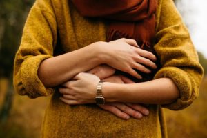 A woman wearing a mustard coloured jumper is hold the the hands of someone who is hugging her from behind. There is no head, just the torso.