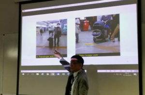 Yoshito Dobashi pointing to his slide at the UDHEIT conference showing wheelchair crossing points, one with a man wheeling a suitcase.