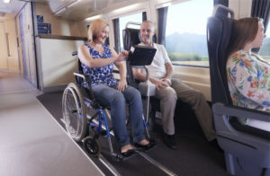 In the train carriage, a woman is seated in a manual wheelchair and is sitting next to a man in a standard seat. They are looking at an in-seat screen, probably for movies.