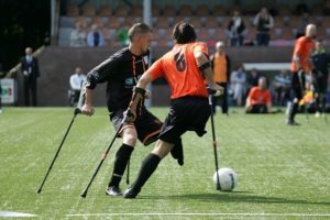 Two young men each with one leg and using crutches, compete for the football on the football field. Kicking UD Goals in Sport. 