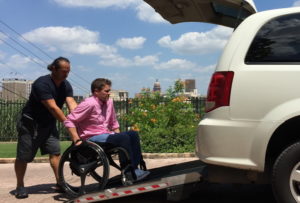 Uber driver is pushing a man in a manual wheelchair into the back of the vehicle.