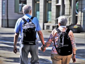 An older man and woman are walking away from the camera down a street. They are wearing backpacks and holding hands.