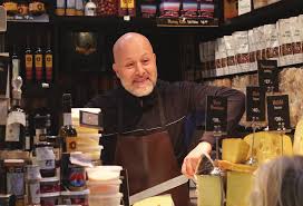 A bald man is standing behind a shop counter that has cheese and other deli items.. More insights and less afterthoughts.