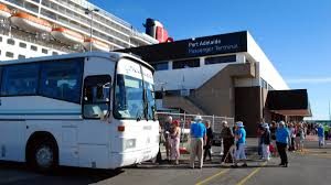 A line pf people are boarding a coach outside a transit building.There is a cruise ship in the backgound.
