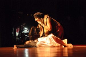 Two women are on stage. One is lying down and looks dead. The other leans over her with grief.