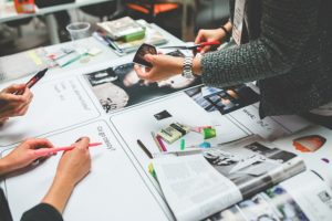 A work table is filled with paper and folders and a woman is cutting a piece of paper with scissors. It looks like a group of people are working on a design.