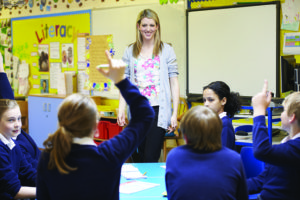 A female teacher stands smiling in front of a class of young students in school uniform. One has her arm raised as if to ask or answer a question.