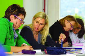 Two pairs of women sit at a table with paper and pens. One of the pair looks to be explaining something to the other. Universal Design for Learning.