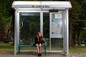 A young woman is sitting in a bus shelter and looking down the road. The shelter is lit and has an information board. Bus and tram stops by universal design.