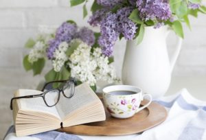 A vase of purple and white flowers sits on a small table with a cup of tea, an open book and a pair of glasses.