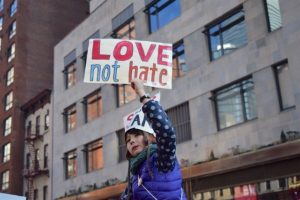 A girl holds up a sign that says, Love not Hate. She appears to be sitting on the shoulders of someone in a public street march.