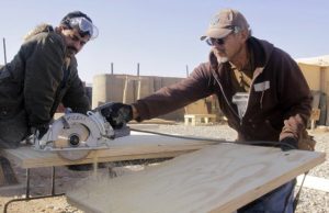 Two men are working on a construction site. One is holding a circular saw which has just cut through a large timber board.