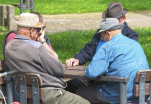 Four older men wearing hats sit at a square table in the park.