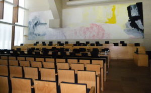 This new courtroom has timber backed seats and a long timber desk that seats the justices. A abstract painting covers the wall behind the bench. Daylight comes in through large windows.