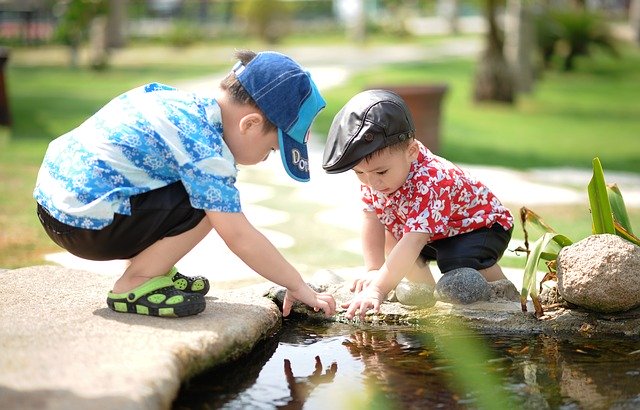 Two small boys are crouched by the side of a pond and are reaching into the water.