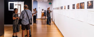 An display space at QUT Art Museum. People are looking at small pictures hanging on a white wall.