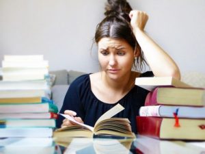 A young woman is sitting with piles of books and is frowning. 