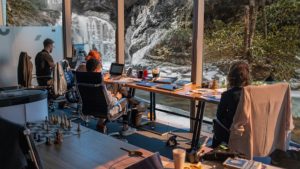 A workbench with computers overlooks a waterfall and stream. People are looking out of the window. Biophilic design is for everyone.